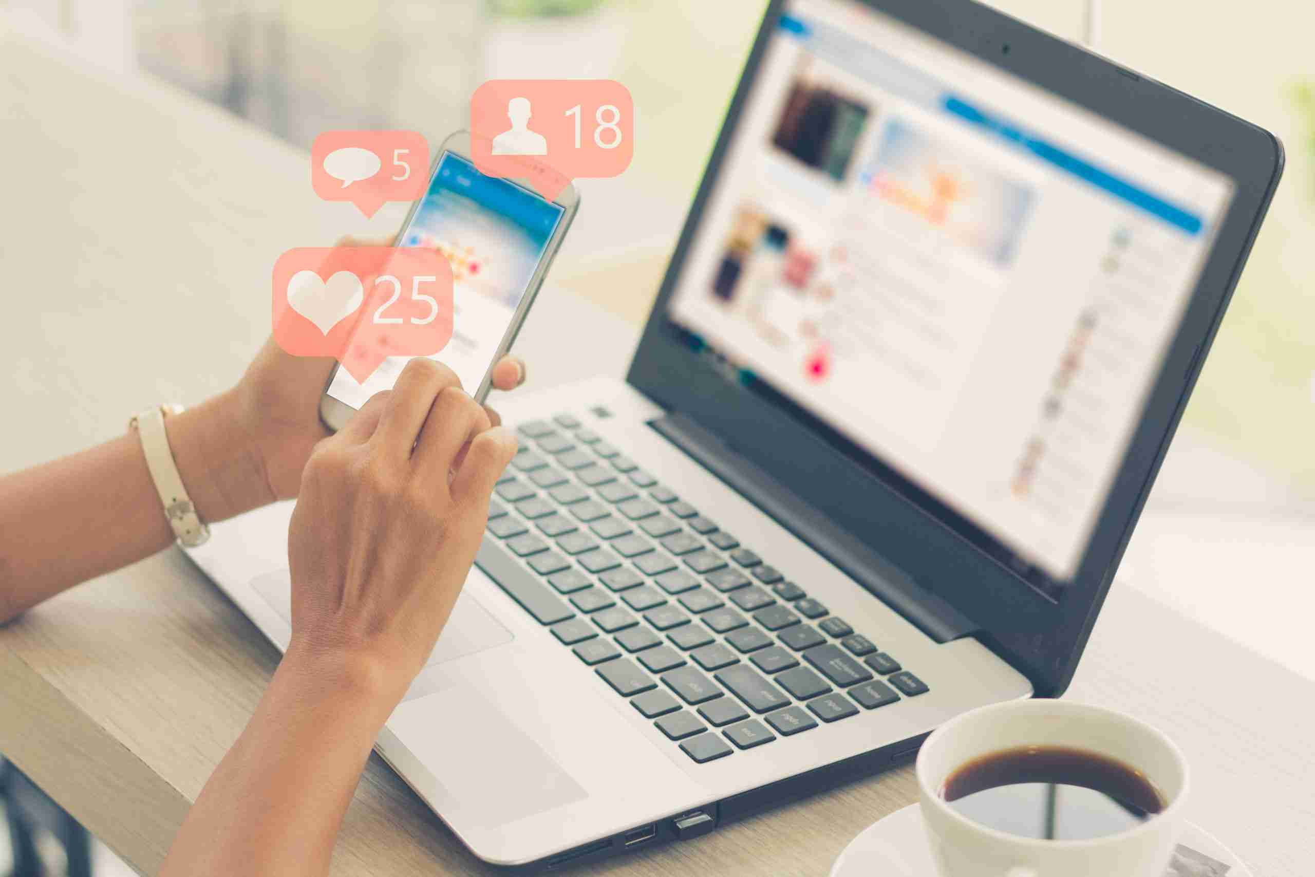 Person using a smartphone with social media notifications next to a laptop on a white table. A cup of coffee sits beside the laptop.