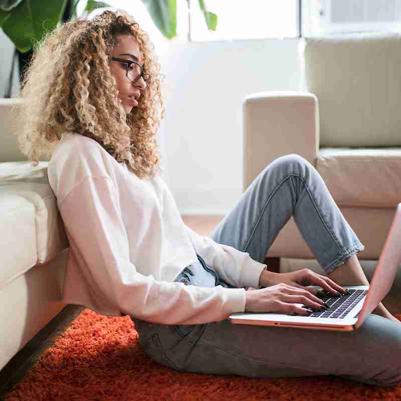 Person with curly hair and glasses sits on the floor, leaning against a couch, typing on a laptop.