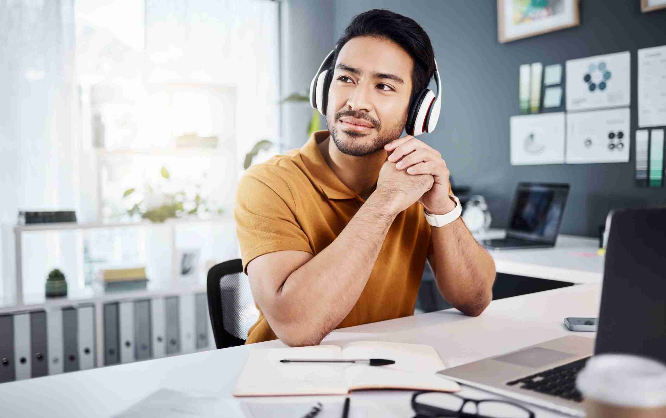 A person wearing headphones sits at a desk, looking thoughtfully to the side. There's an open notebook, a laptop, and various charts on the wall in the background.